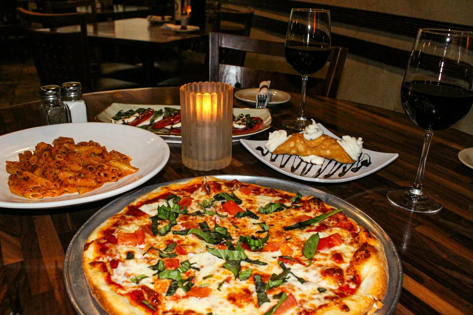 Pizza med tomat og basilikum, pasta og to glas rødvin på et restaurantbord